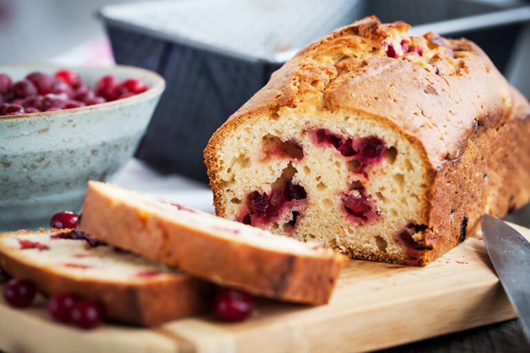 Sliced cranberry loaf on a cutting board beside a bowl of cranberries.