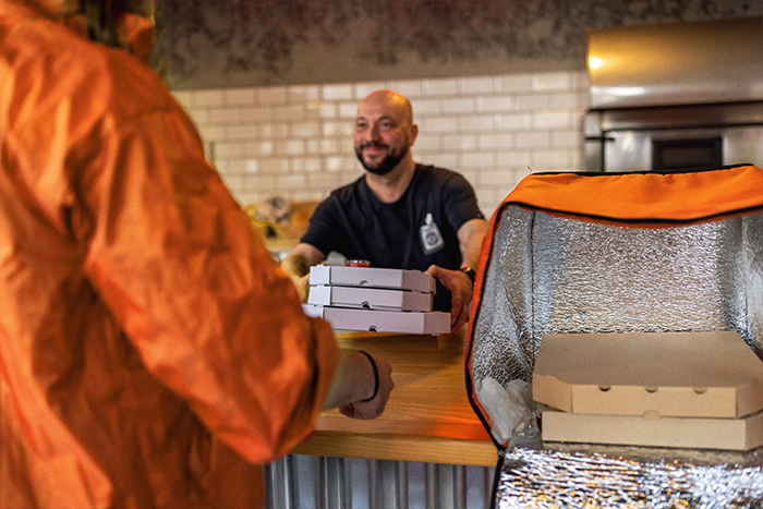 A chef handing three boxes over a counter to a delivery person with an insulated bag open beside them.