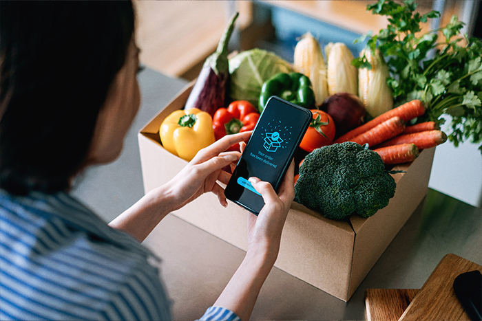 Person holding their phone receiving an order in front of a box of fresh vegetables.