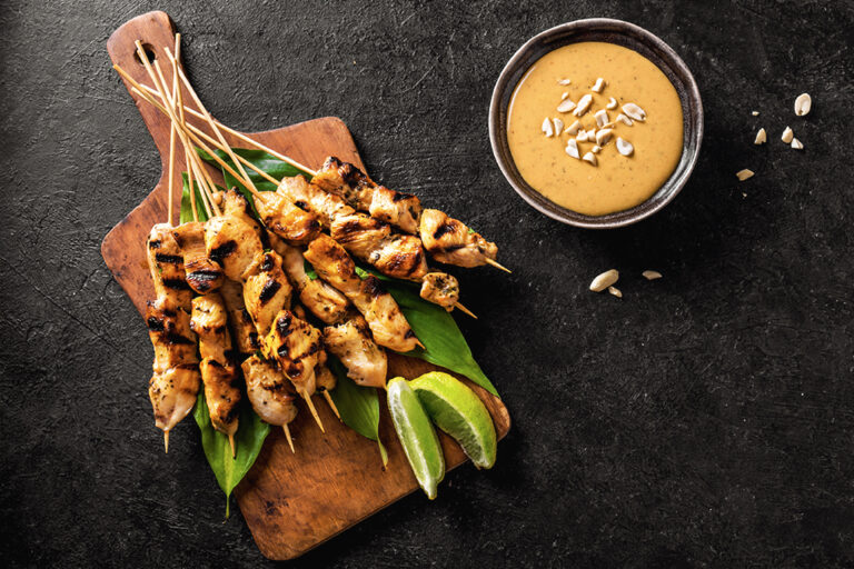 Chicken skewers served on a wood board with lime wedges, paired with a ramekin of peanut sauce.