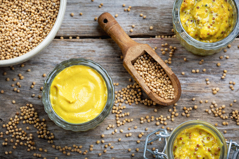Three glass bowls of prepared mustard and a bowl of mustard seeds on a wooden table.